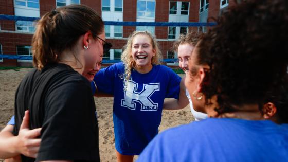 Laughing students with K Crew leader huddled on sand volleyball court